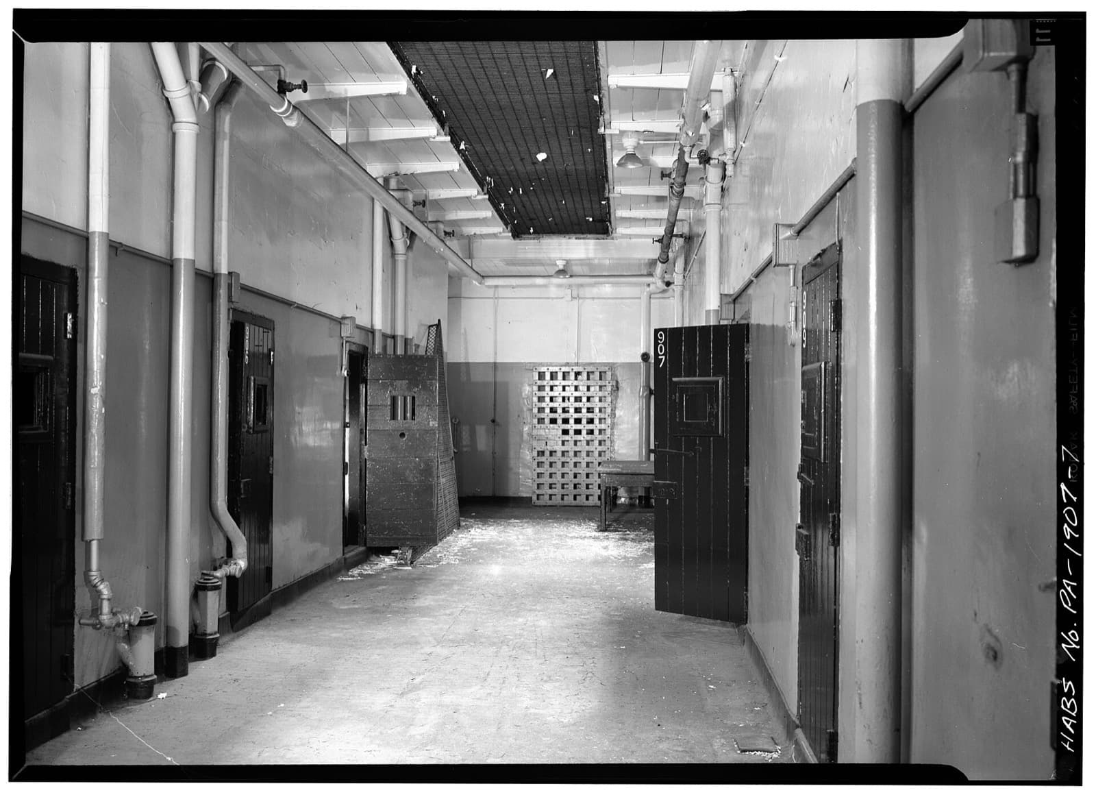 A corridor of cells in a historic prison, in black-and-white archive photography: numbered iron doors, exposed pipework and a concrete floor