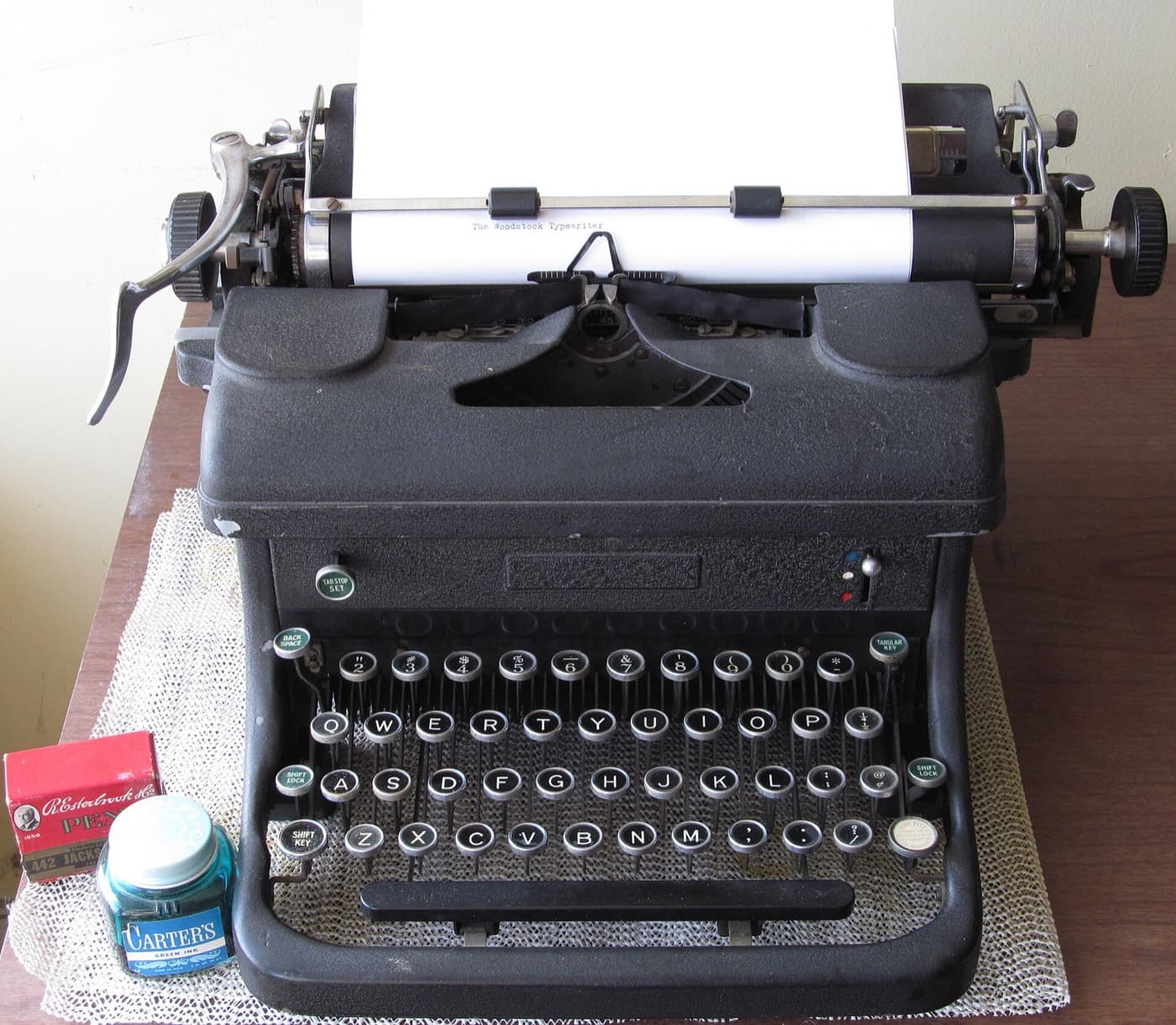 A 1940s Woodstock typewriter with a sheet of paper in the platen; beside it an ink bottle and a box of pens